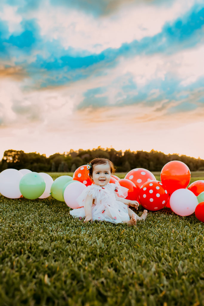 infant baby girl celebrating first birthday with a photoshoot in a field with balloons