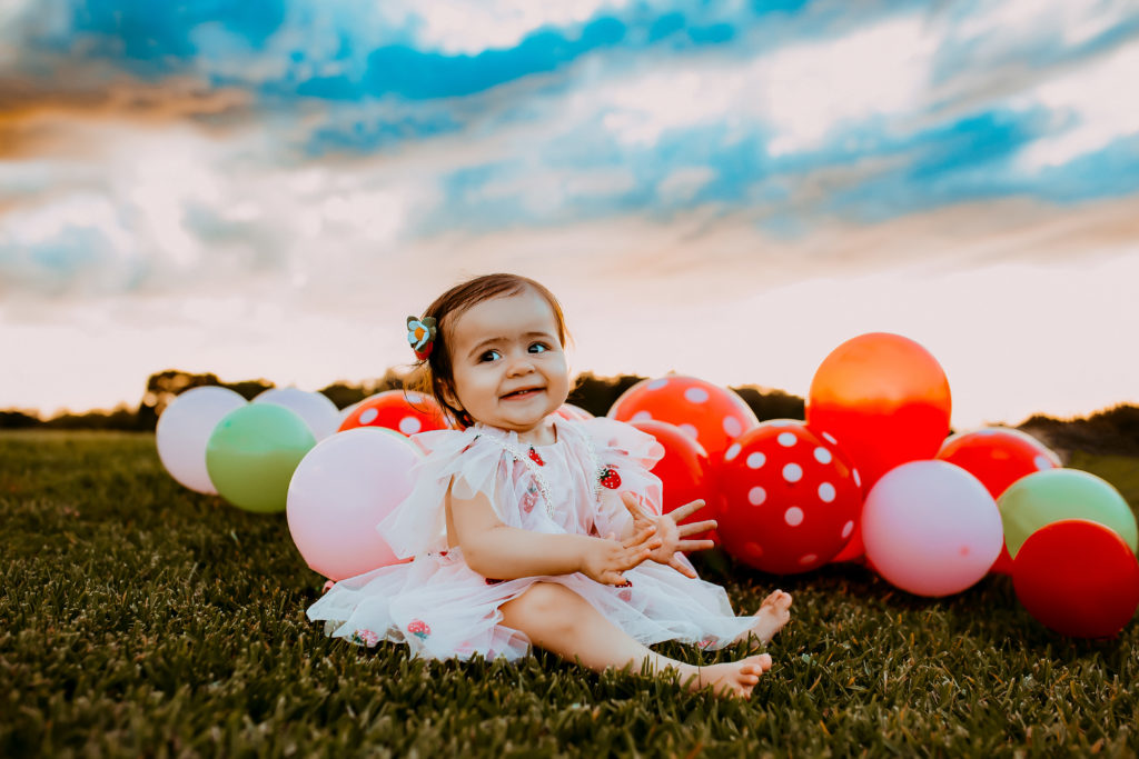 one year old wearing a tulle strawberry dress sitting in a field at sunset with balloons in the background