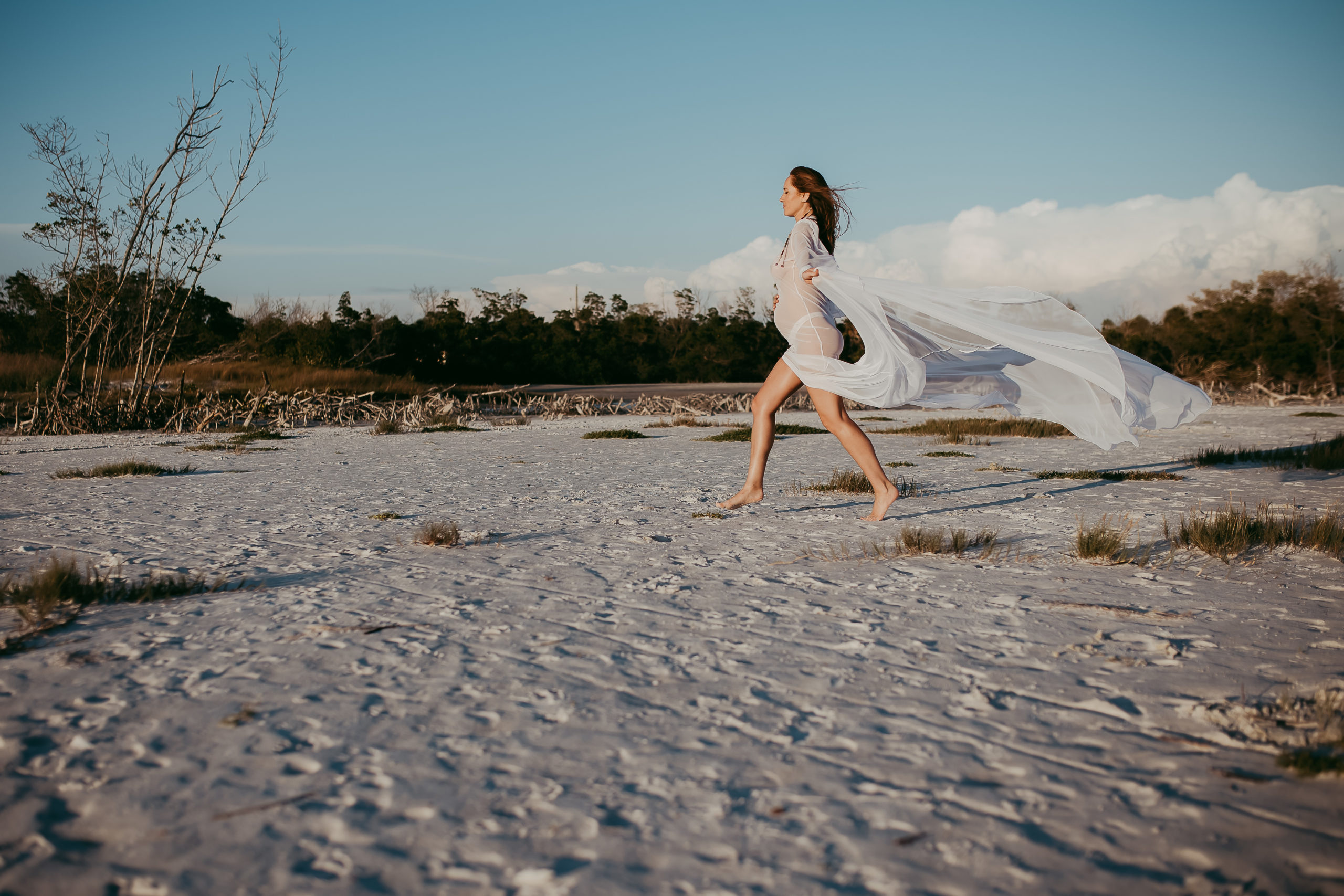 maternity beach photoshoot of mother running on the beach holding the long train on her white sheer dress