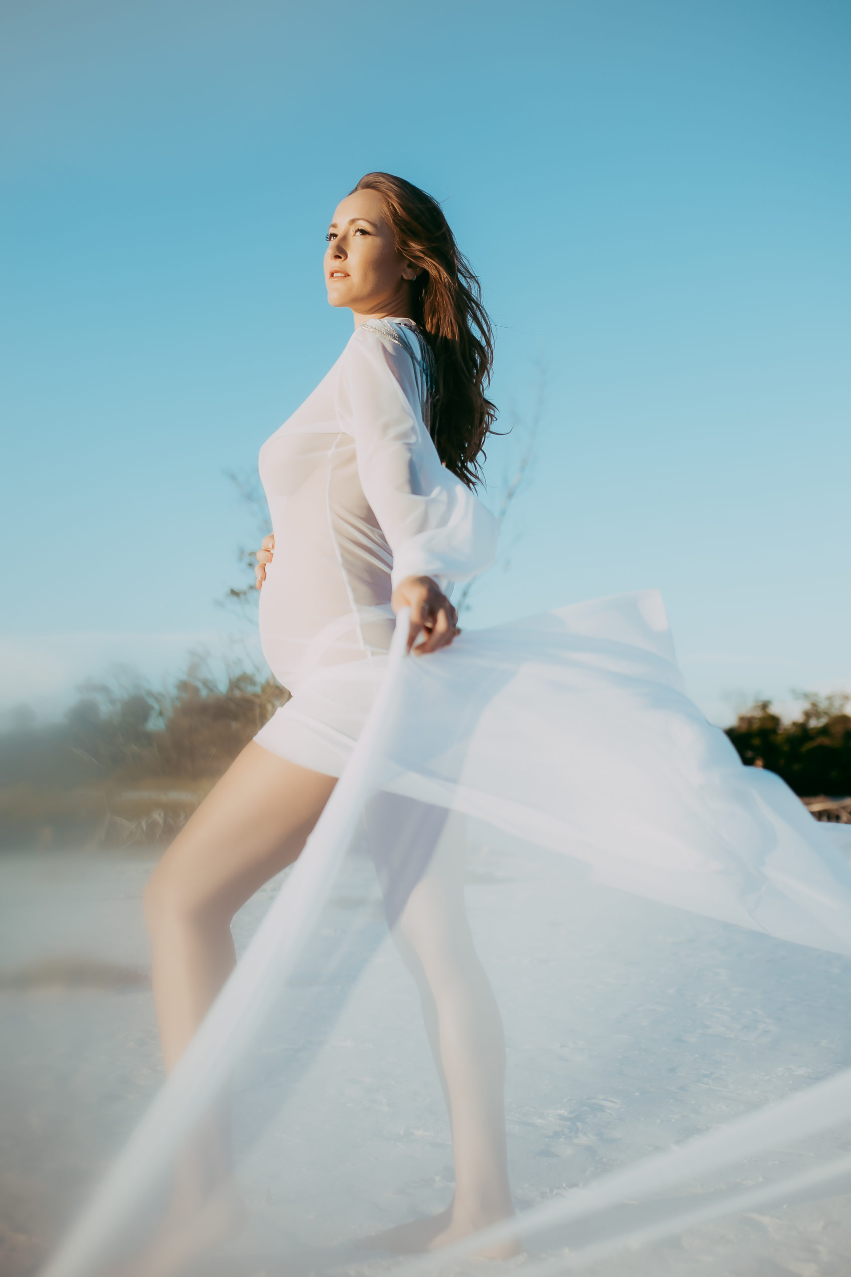 ethereal beach maternity outdoor photoshoot of pregnant mom waving her long sheer dress around glancing toward the ocean at the beach and holding her baby bump