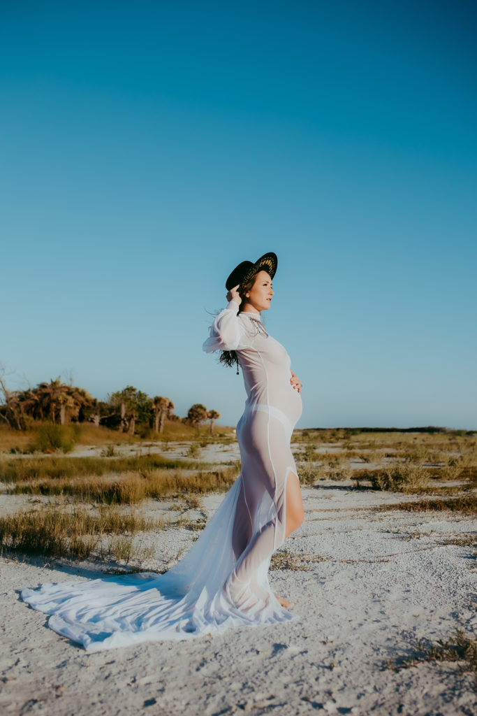Bohemian style beach maternity photoshoot where the pregnant mother is wearing a turtle neck long sleeve white sheer dress with a long train and wearing a black hat