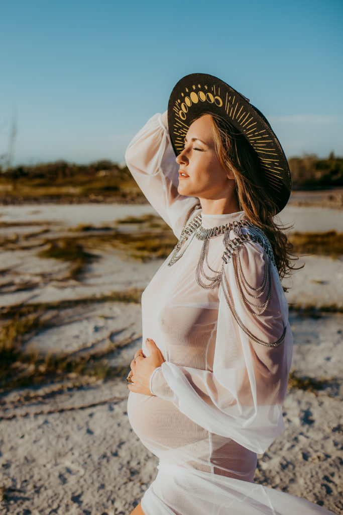 Pregnant mother holding her belly and standing on the beach with the direct harsh sun hitting her and she is wearing a sheer see through dress and chain shoulder piece