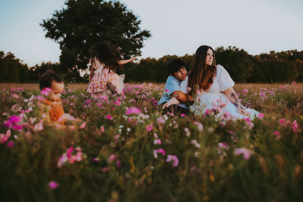 Outdoor motherhood photoshoot in a field of wild purple flowers. A mother is sitting in the tall grass with her oldest child hugging her and the other two toddlers running around and exploring.