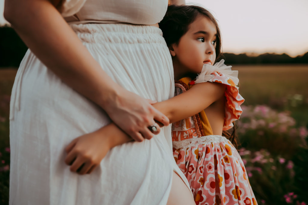 A young girl hugging her mom tightly around her waist and looking out at the sun setting over the field of wildflowers