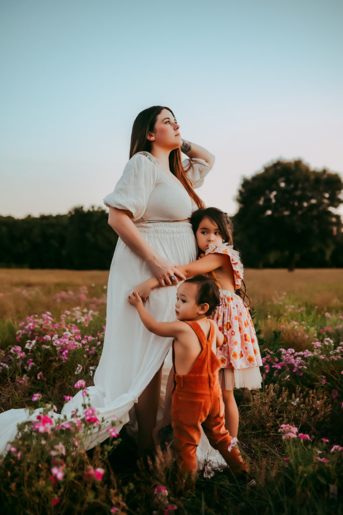 A mom standing in a large open field of wildflowers as her two young children hug around her tightly for an outdoor motherhood photoshoot