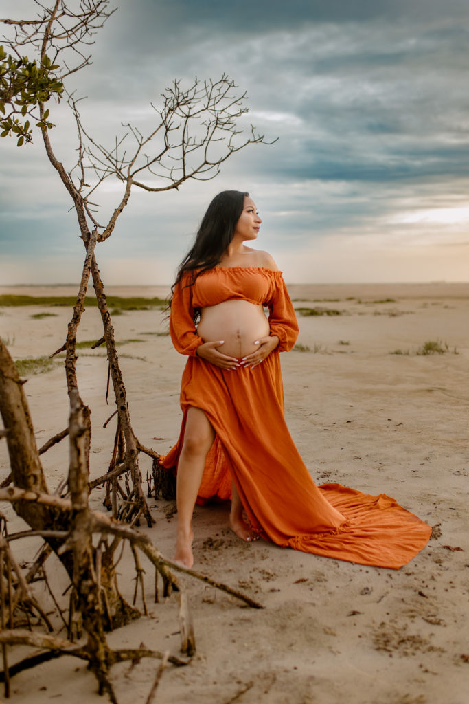 pregnant mother wearing an orange two piece maternity gown with her bare belly out. she is standing next to trees with exposed roots on the beach for a maternity photoshoot