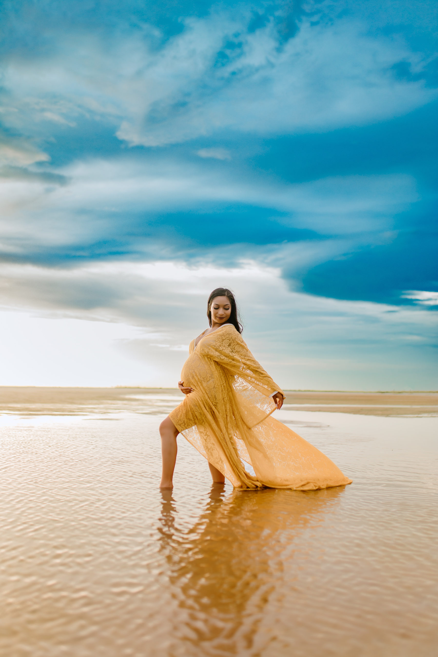 gorgeous pregnant mom standing in the water against a vivid blue cloudy sky with her reflection in the water for a beach maternity photoshoot