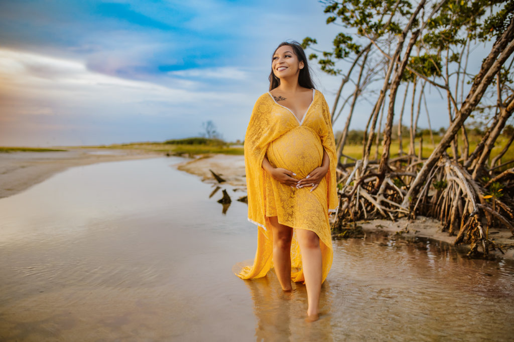 maternity beach photo session with pregnant mom standing in the water wearing a yellow lace dress holding her baby bump