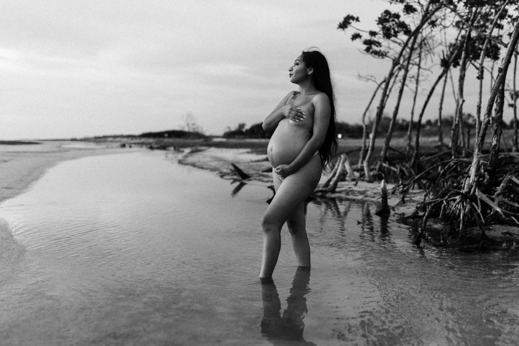 black and white nude maternity portrait of mother holding her baby bump standing in a river of water at the beach during sunset