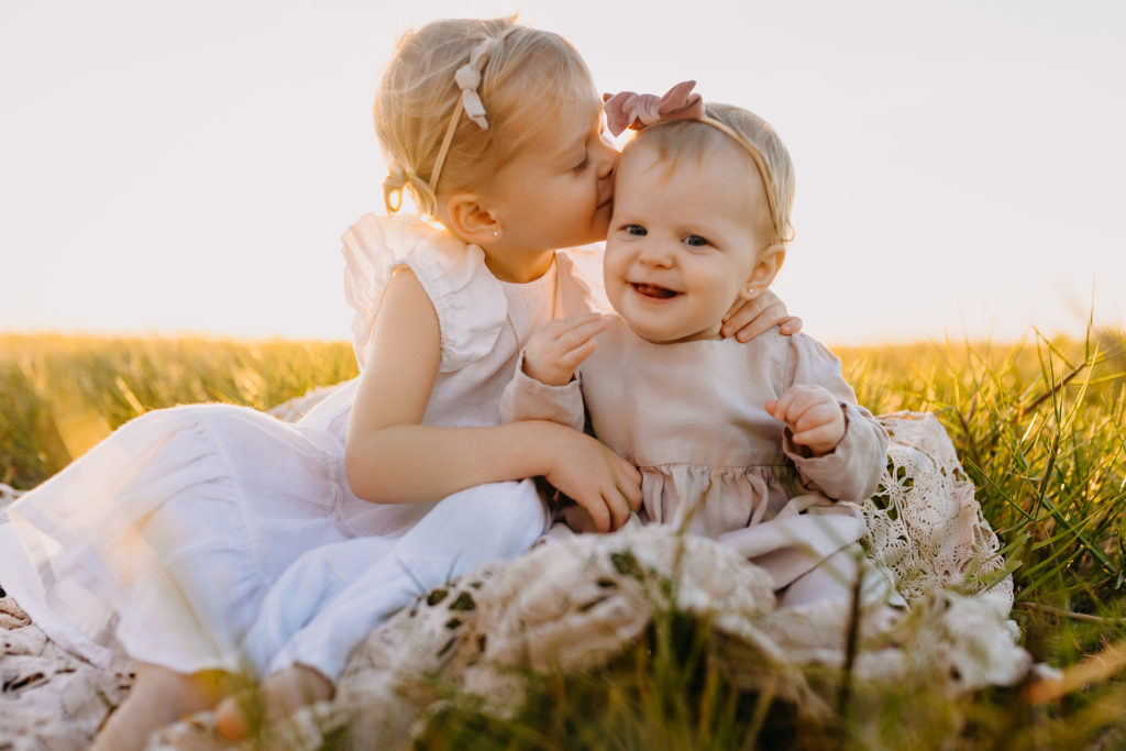 Three year old toddler girl wearing a white dress sitting next to her 12 month old sister giving her sweet kisses on her cheek