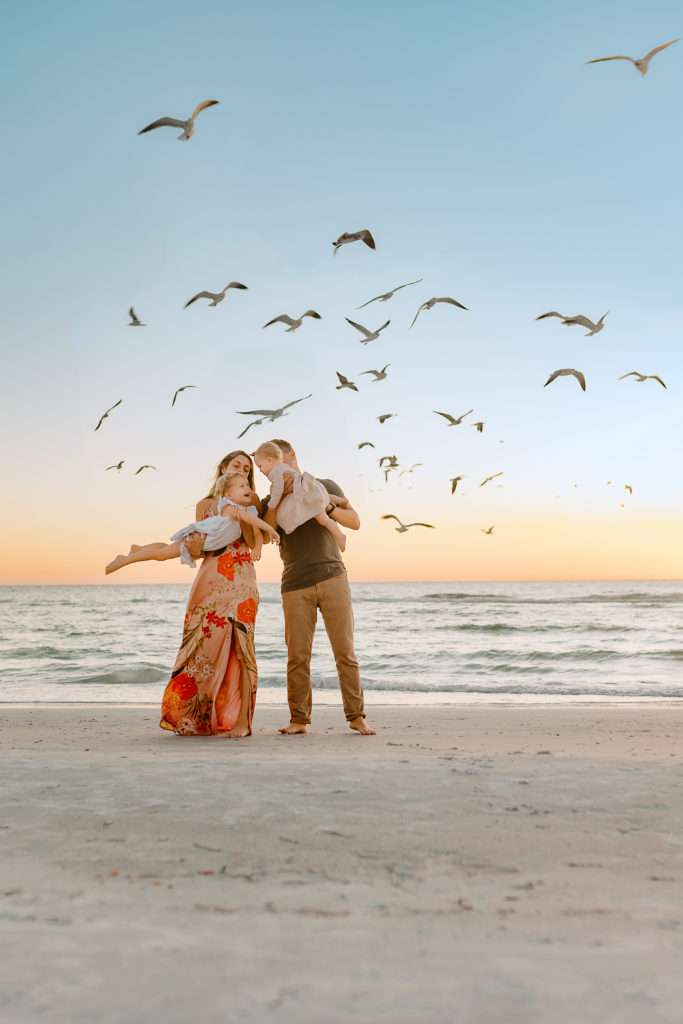 Sunset beach photo with seagulls flying in the background of mom and dad standing holding the children like airplanes flying them around playfully