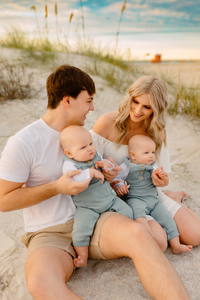 mom and dad sitting in front of sea grass on the beach holding their twin baby boys