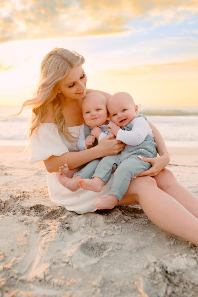 mother and her twin baby boys sitting in her lap as they are holding one another and smiling for family beach portraits
